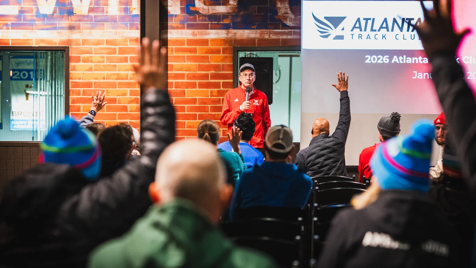 Man in red jacket speaking to an audience raising hands at an Atlanta Track Club event indoors.