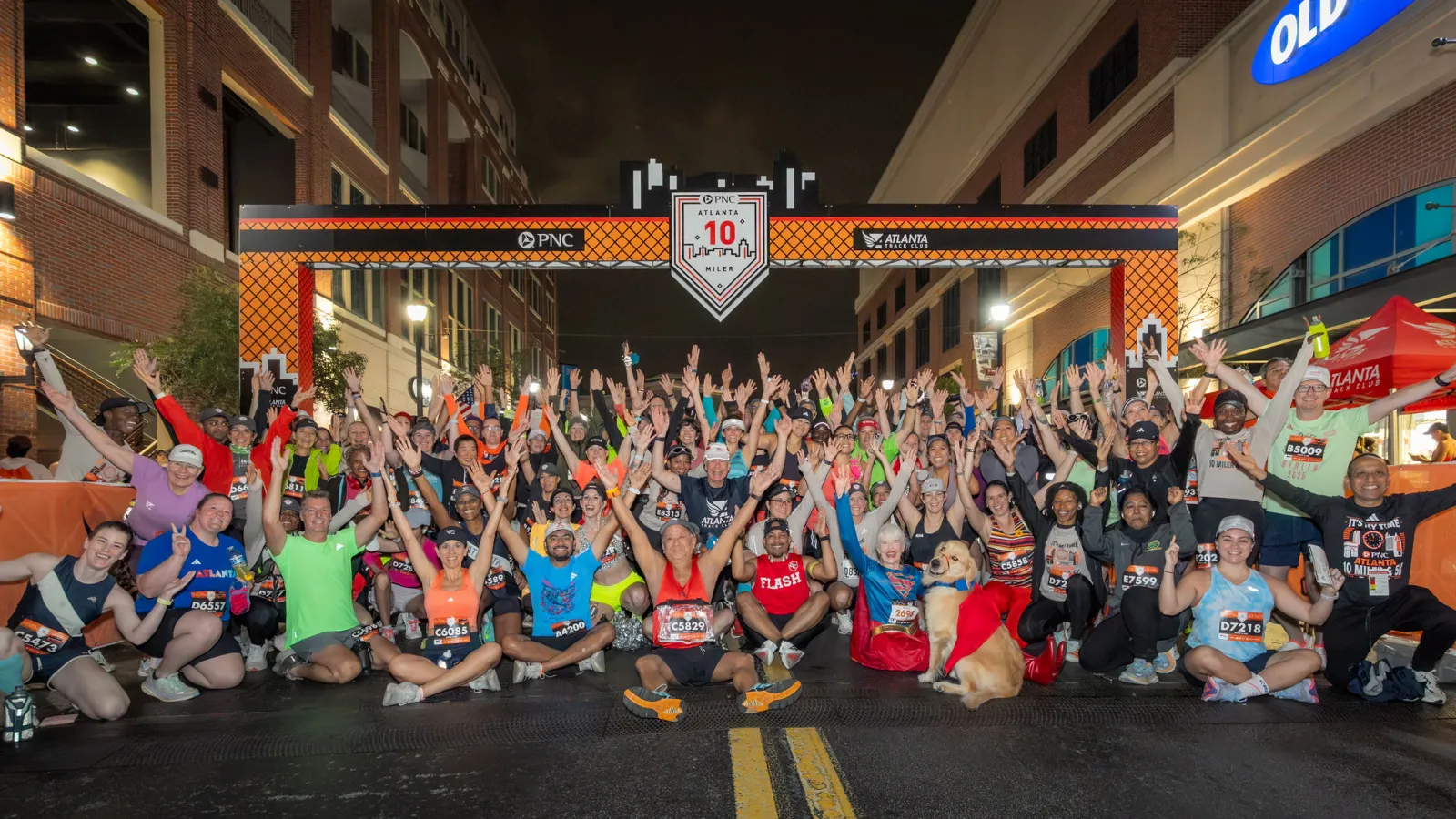 Large group of runners celebrating under a race finish line banner at night on a city street.