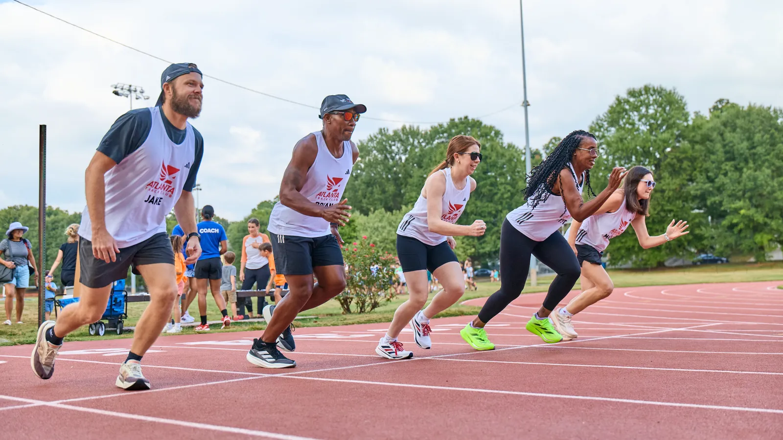 Diverse group of five runners starting a race on a red track under a cloudy sky with green trees in the background
