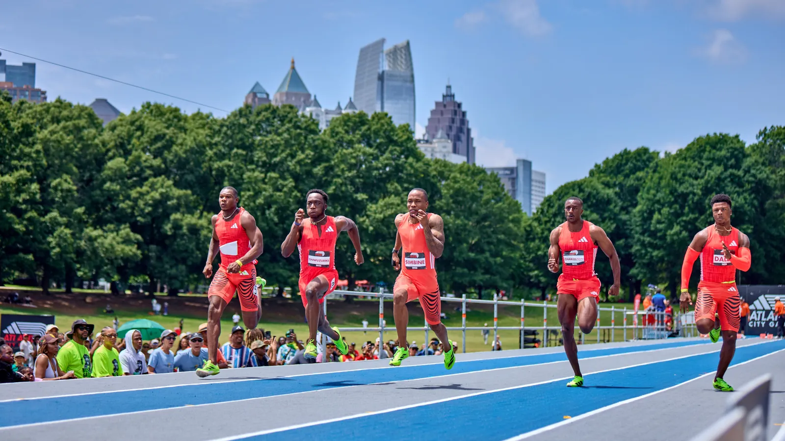 Five male athletes sprinting on a blue track with a city skyline and green trees in the background under a blue sky.