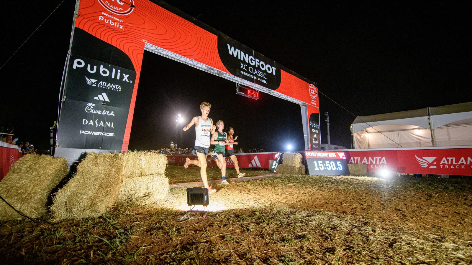 Runners cross finish line at night under Wingfoot XC Classic arch with event sponsors and timer visible.