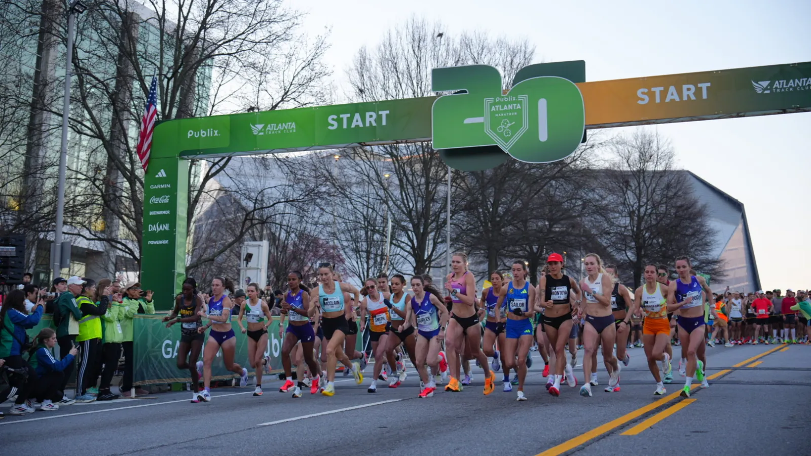 Female runners start race under 20th annual Atlanta marathon green and orange start banner on city street.