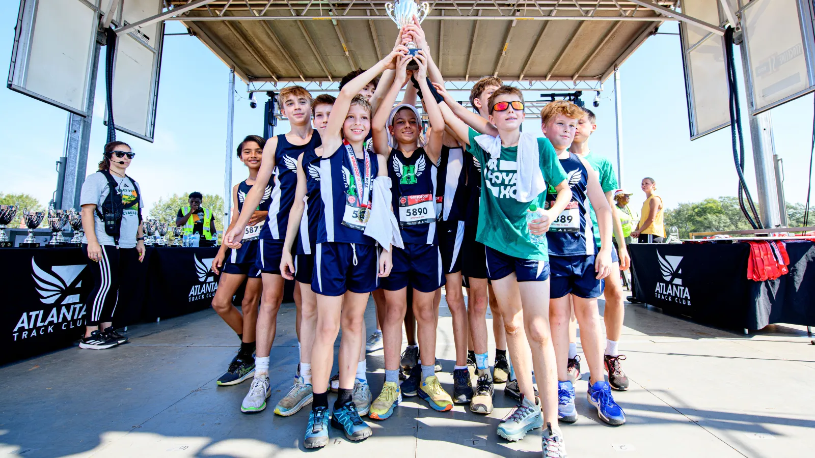 Youth track team in navy uniforms celebrates holding a trophy on an outdoor stage at an Atlanta Track Club event.