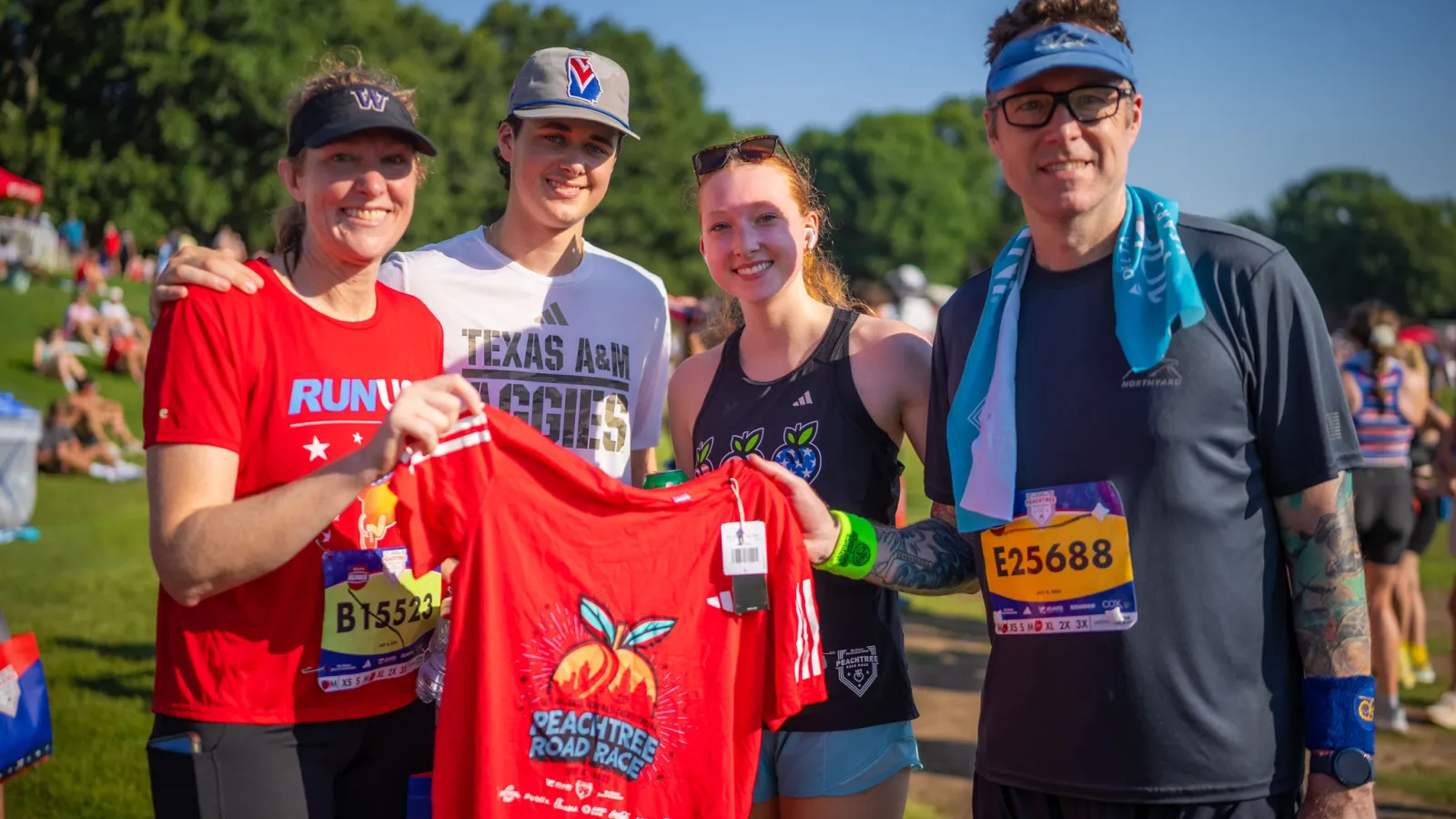 Four smiling runners in race gear holding a red Peachtree Road Race t-shirt outdoors.