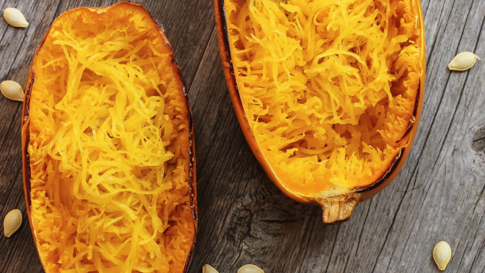 Two halves of a butternut squash are open and facing up towards the camera while on top of an outdoor deck. There are squash seeds sporadically placed around them. The inside of the halves are shredded with a fork which is placed on the top of the right half.