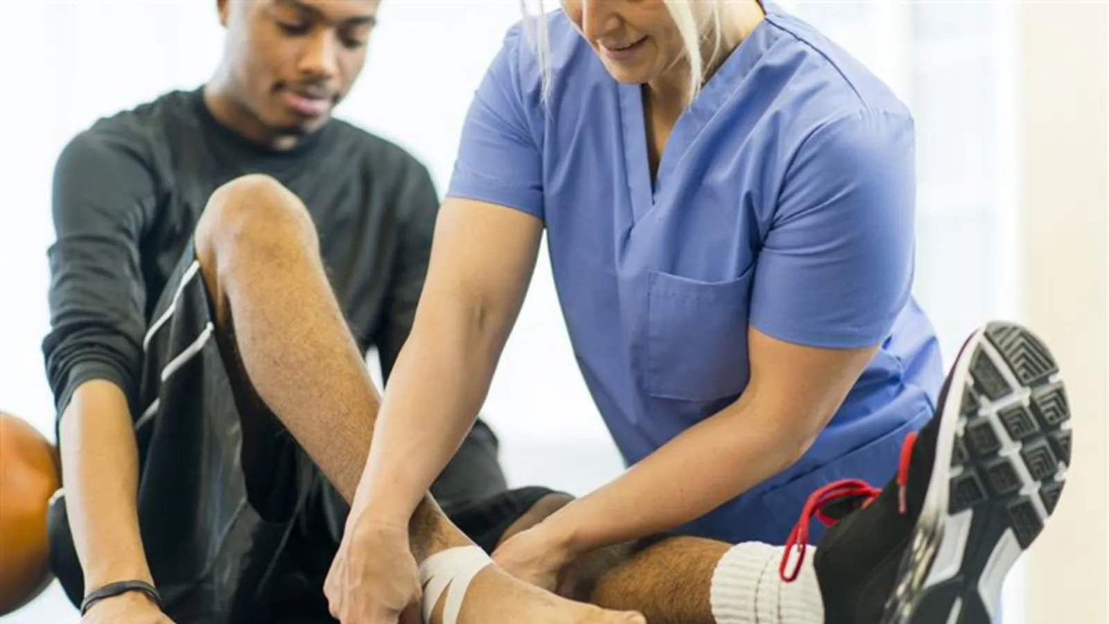 Athletic trainer in blue scrubs wrapping ankle of young male athlete during sports therapy session