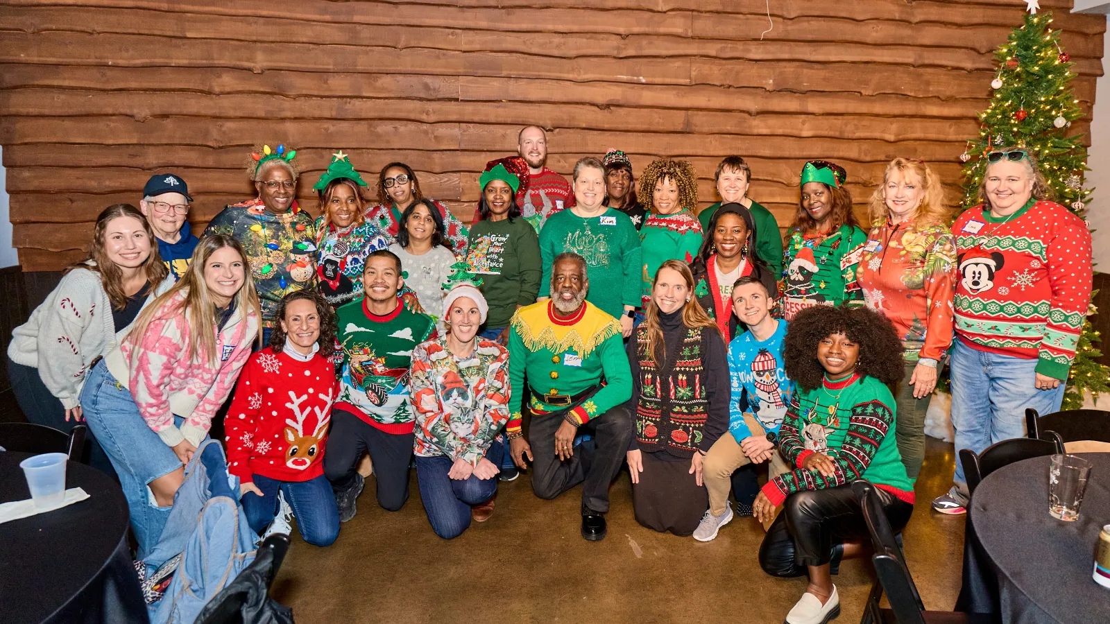 Group of diverse adults in festive holiday sweaters posing indoors near a decorated Christmas tree