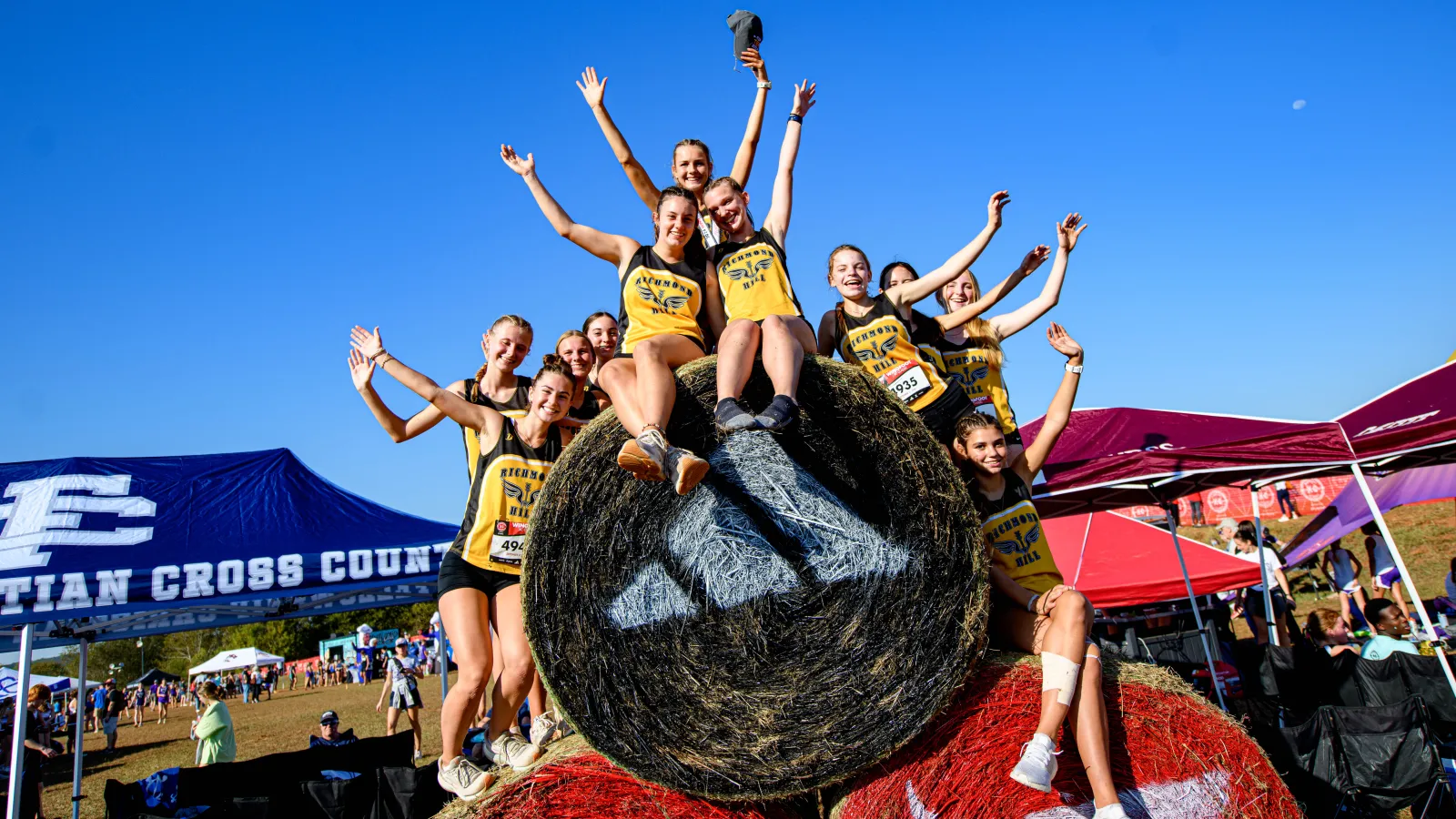 Group of female cross country runners sitting on hay bales, celebrating with arms raised under clear blue sky