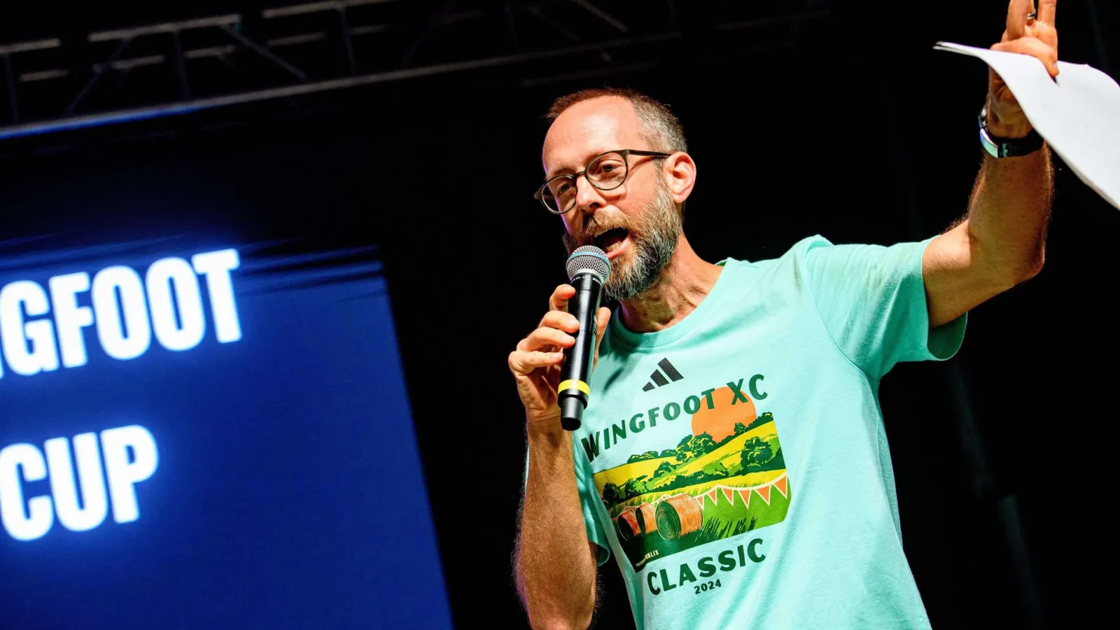 Man with glasses and beard speaking into microphone wearing Wingfoot XC Classic 2024 event t-shirt on stage