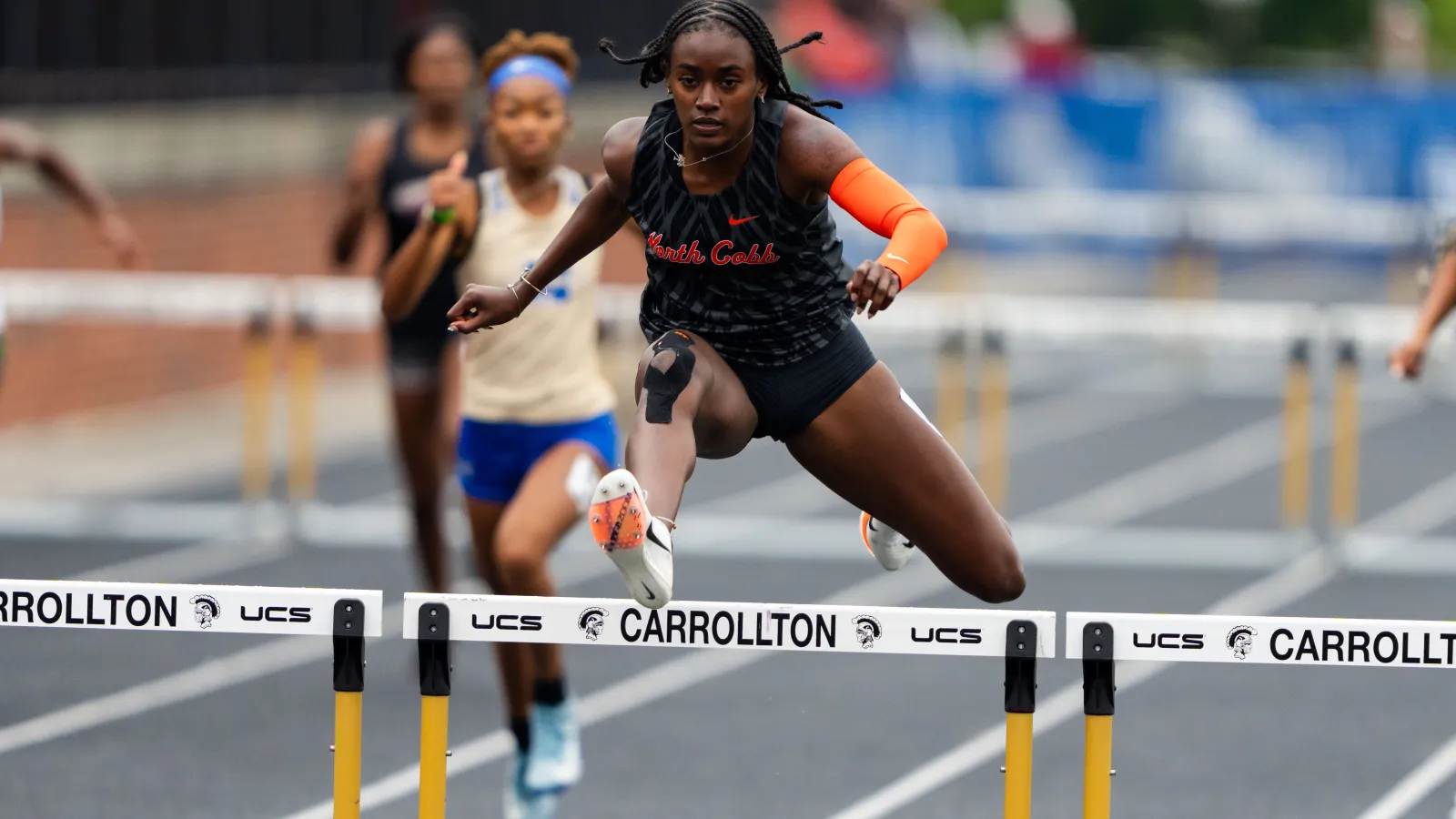 Athlete in black uniform jumping over hurdle during a track and field race with competitors in background