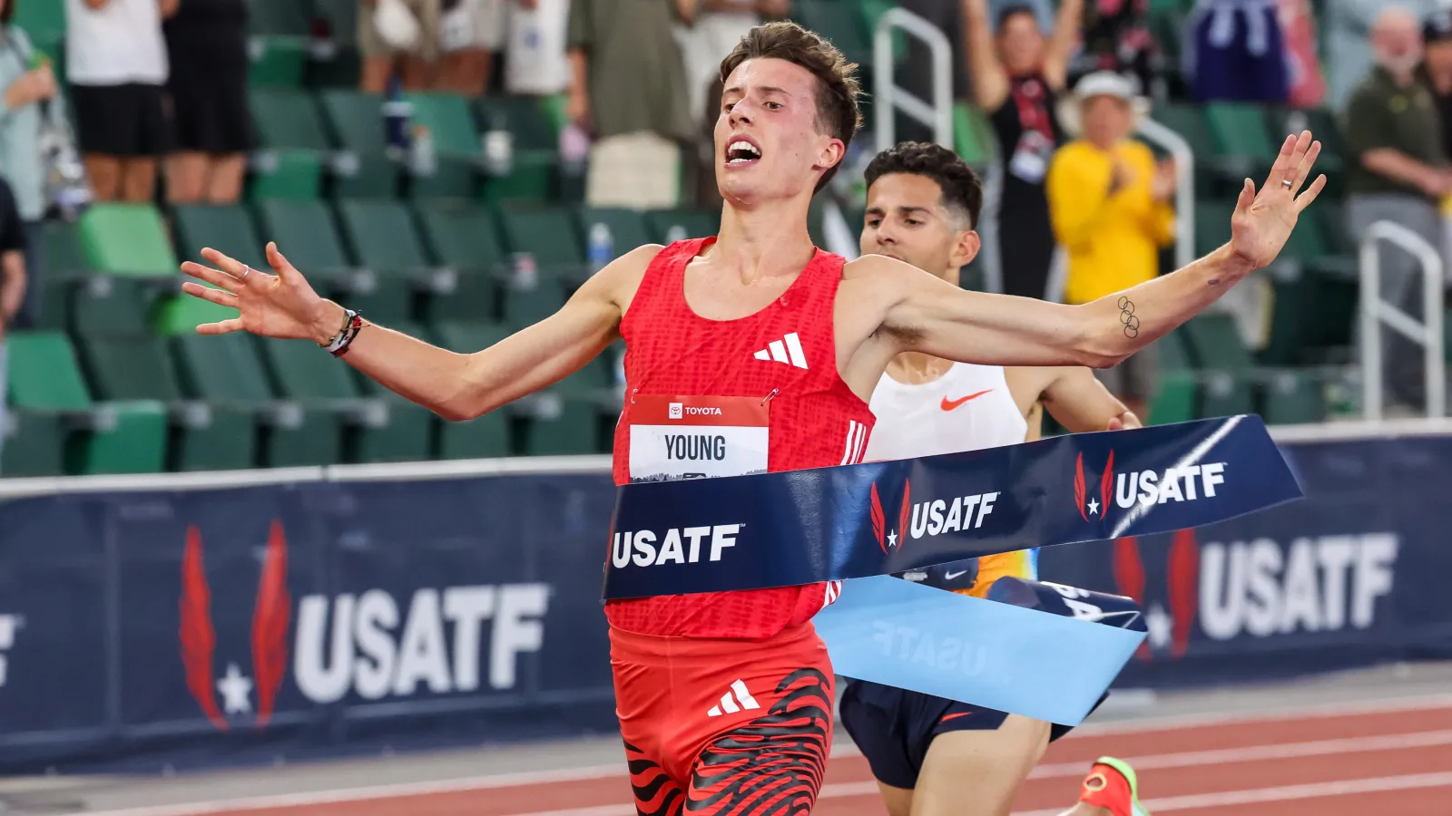 Male runner in red crosses finish line first in USATF track race with competitor behind and crowd cheering