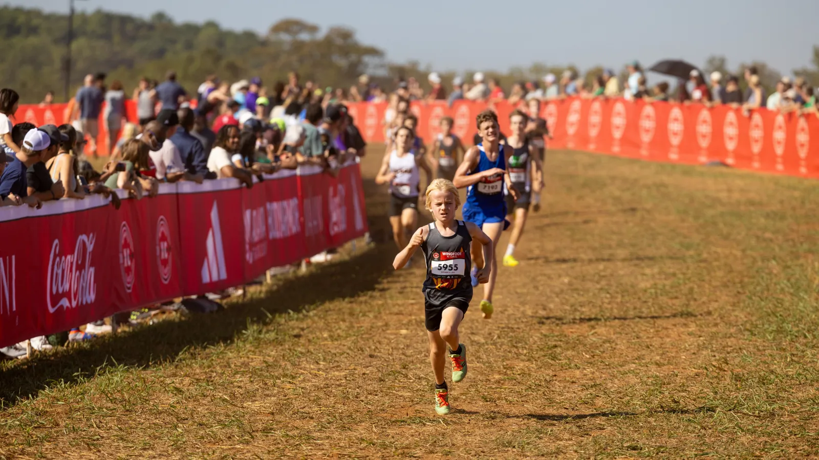 Young athletes running in a cross-country race with spectators cheering along a red-bannered course.