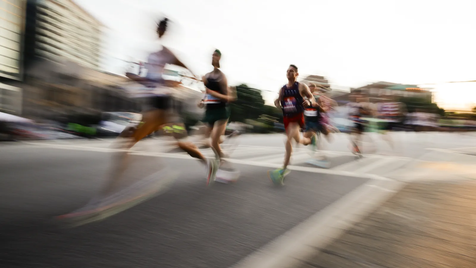 Blurred runners competing in a road race during daytime with city buildings in the background.