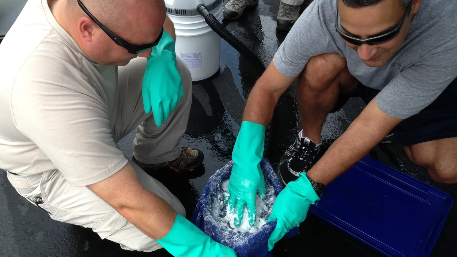 Two men wearing green gloves handle ice or water inside a blue container on a wet surface outdoors.