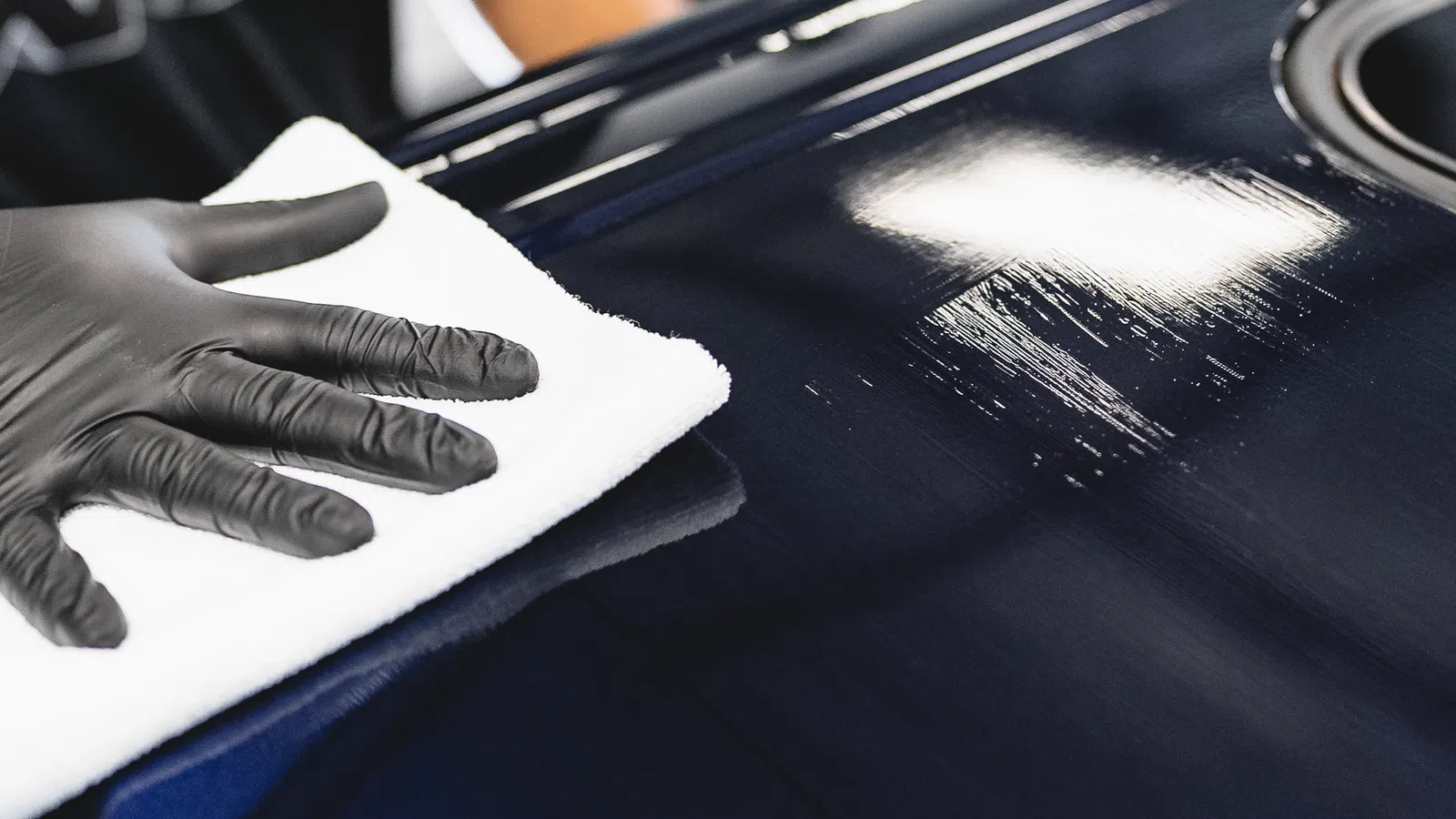 Person wearing black gloves polishing a shiny dark blue car hood with a white cloth indoors.