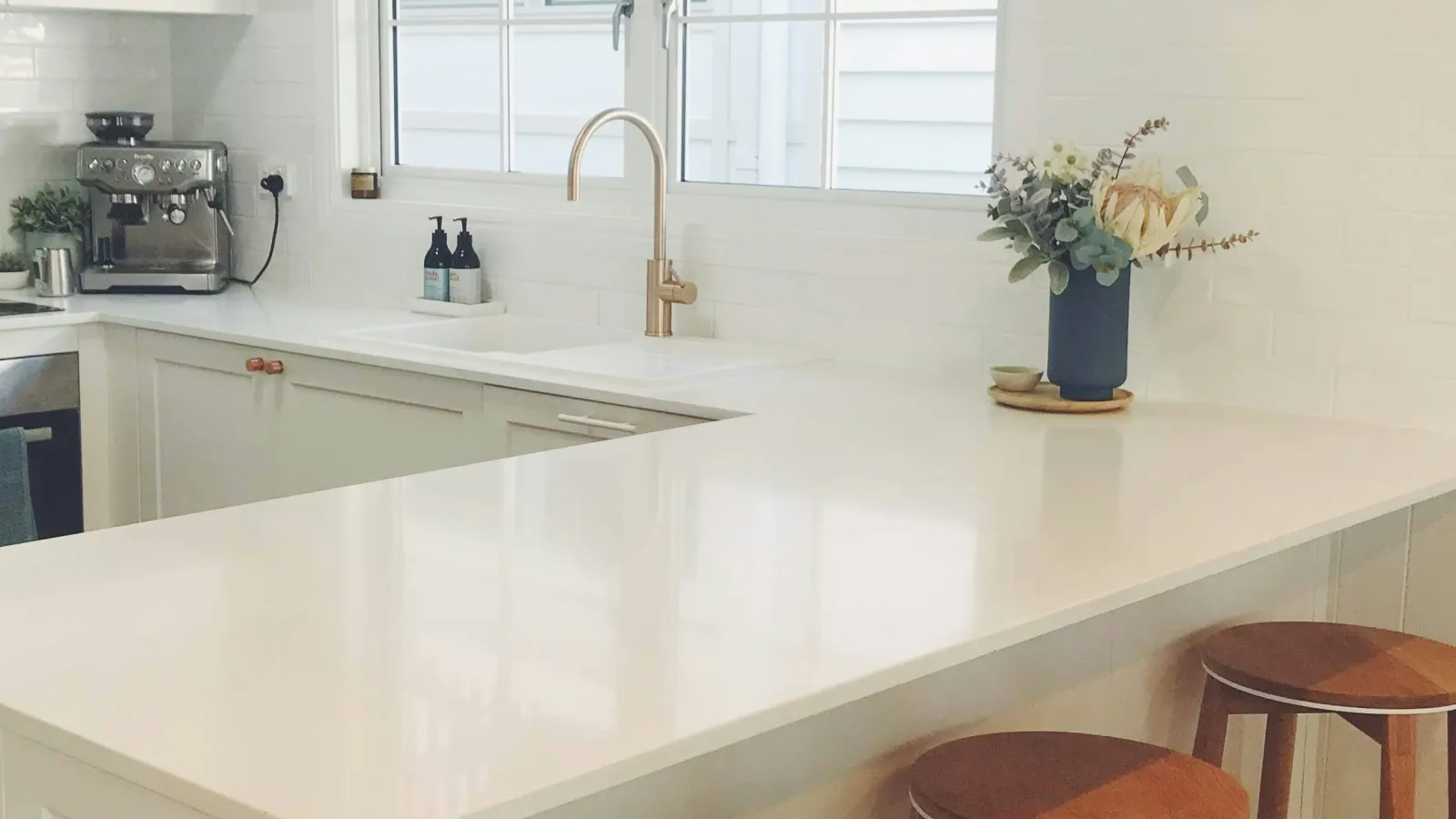 Bright modern kitchen with white countertops, wooden stools, gold faucet, and blue wall clock by window.