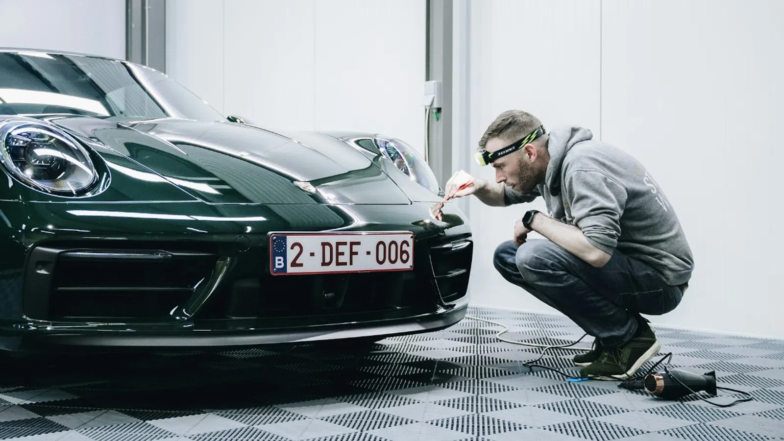 Technician inspecting and polishing a dark green sports car inside a well-lit garage with CarbonQ logo on wall.