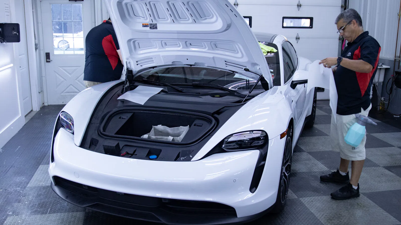 White sports car with open front trunk being inspected by two men inside a garage with checkered floor.