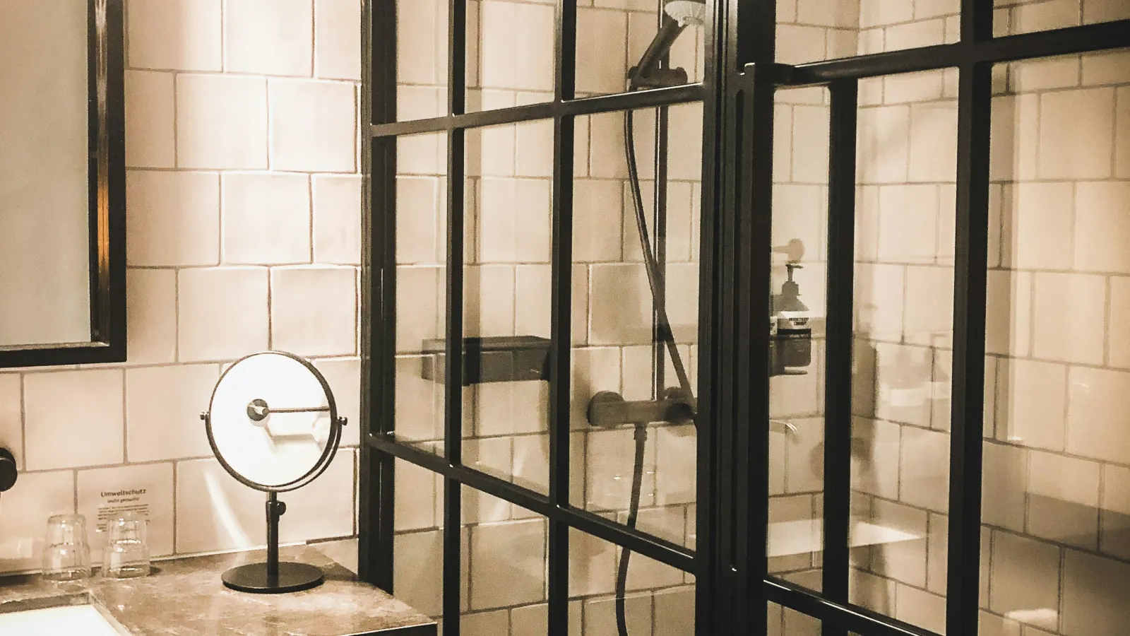 Modern bathroom with black-framed glass shower, white tile walls, dark vanity, and round mirror with towel.