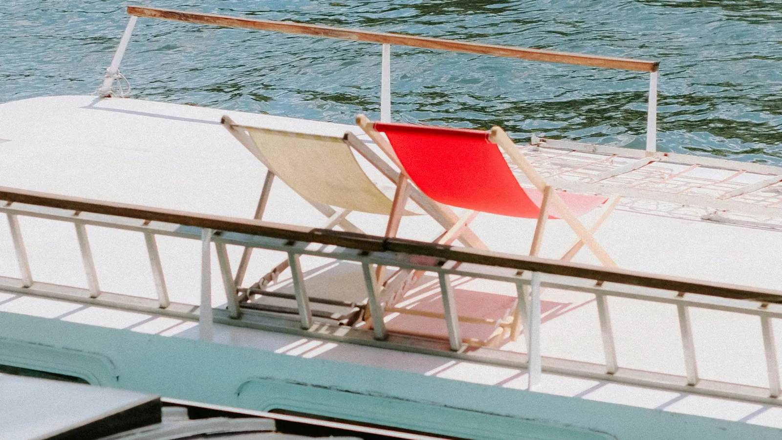 Two deck chairs, one red and one beige, on a boat deck beside calm waterfront under bright sunlight.