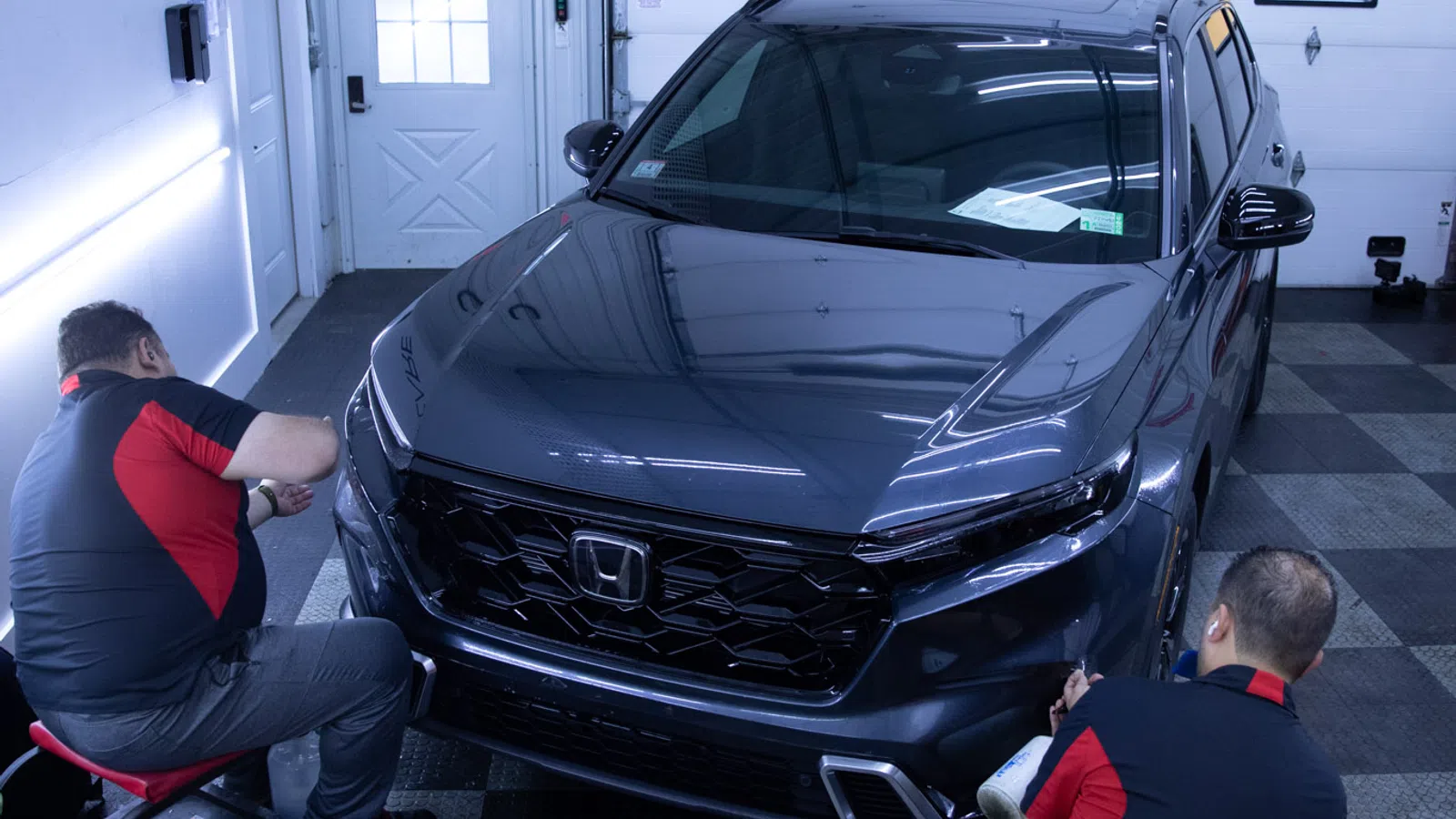 Two mechanics detailing a black Honda SUV inside a garage with checkered flooring and white walls.