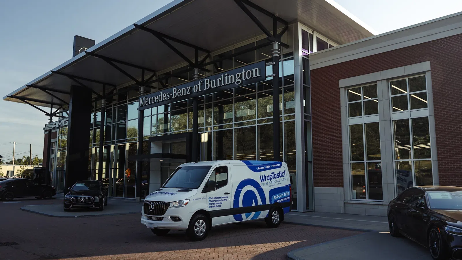 White WrapPlastics van parked in front of Mercedes-Benz of Burlington dealership with luxury cars and modern building