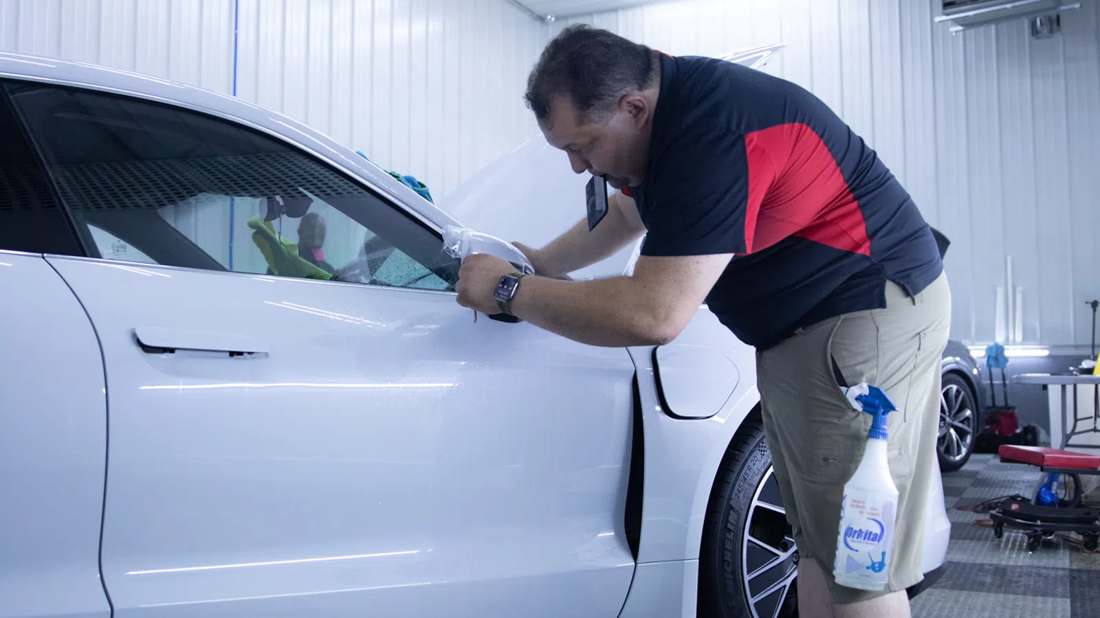 Man detailing a white car's side mirror in a well-lit garage with cleaning supplies on the floor and in his pocket.
