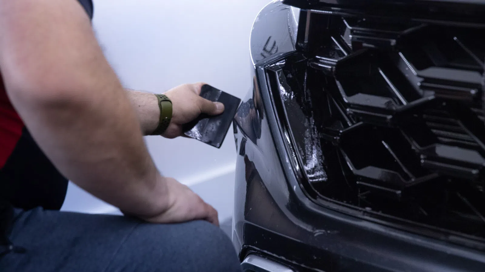 Technician applying vinyl wrap or protective film to the front bumper of a black car in a workshop.