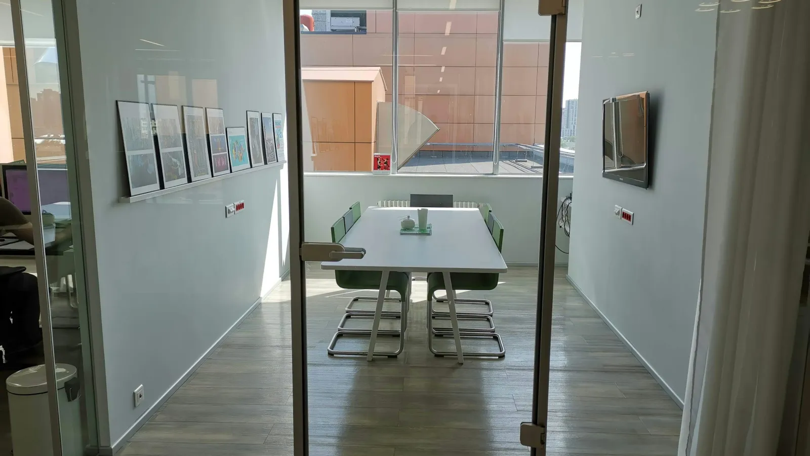 Modern small office meeting room with white table, green chairs, glass walls, and natural light from window.