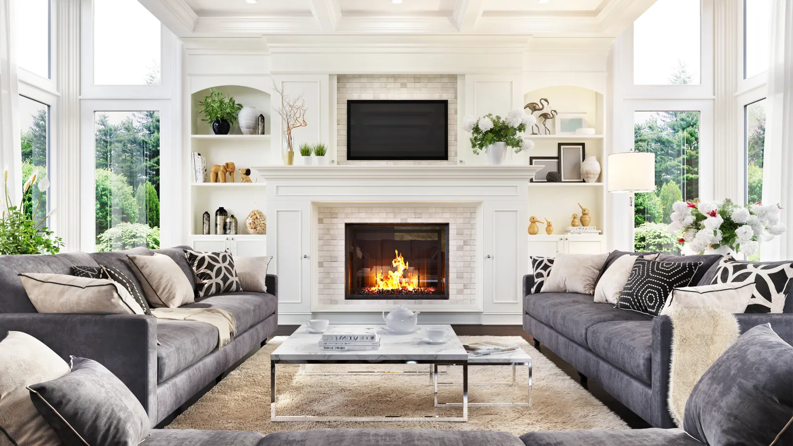 Bright modern living room with gray sofas, a roaring fireplace, large windows, and decorative shelves with plants and art.