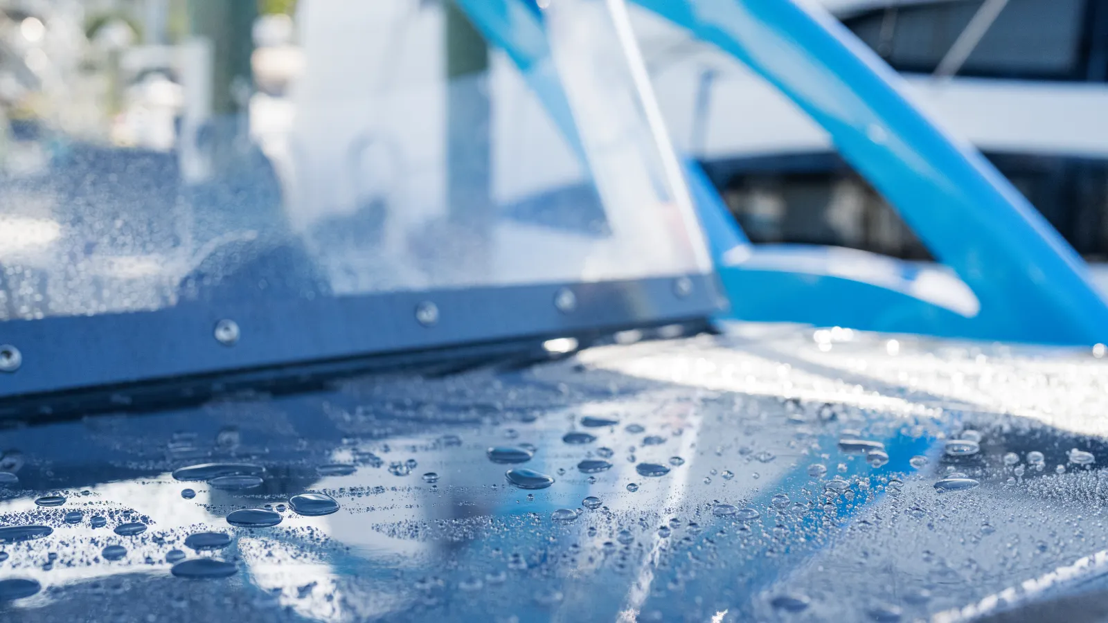 Close-up of water droplets on a blue car's hood and windshield in bright daylight.