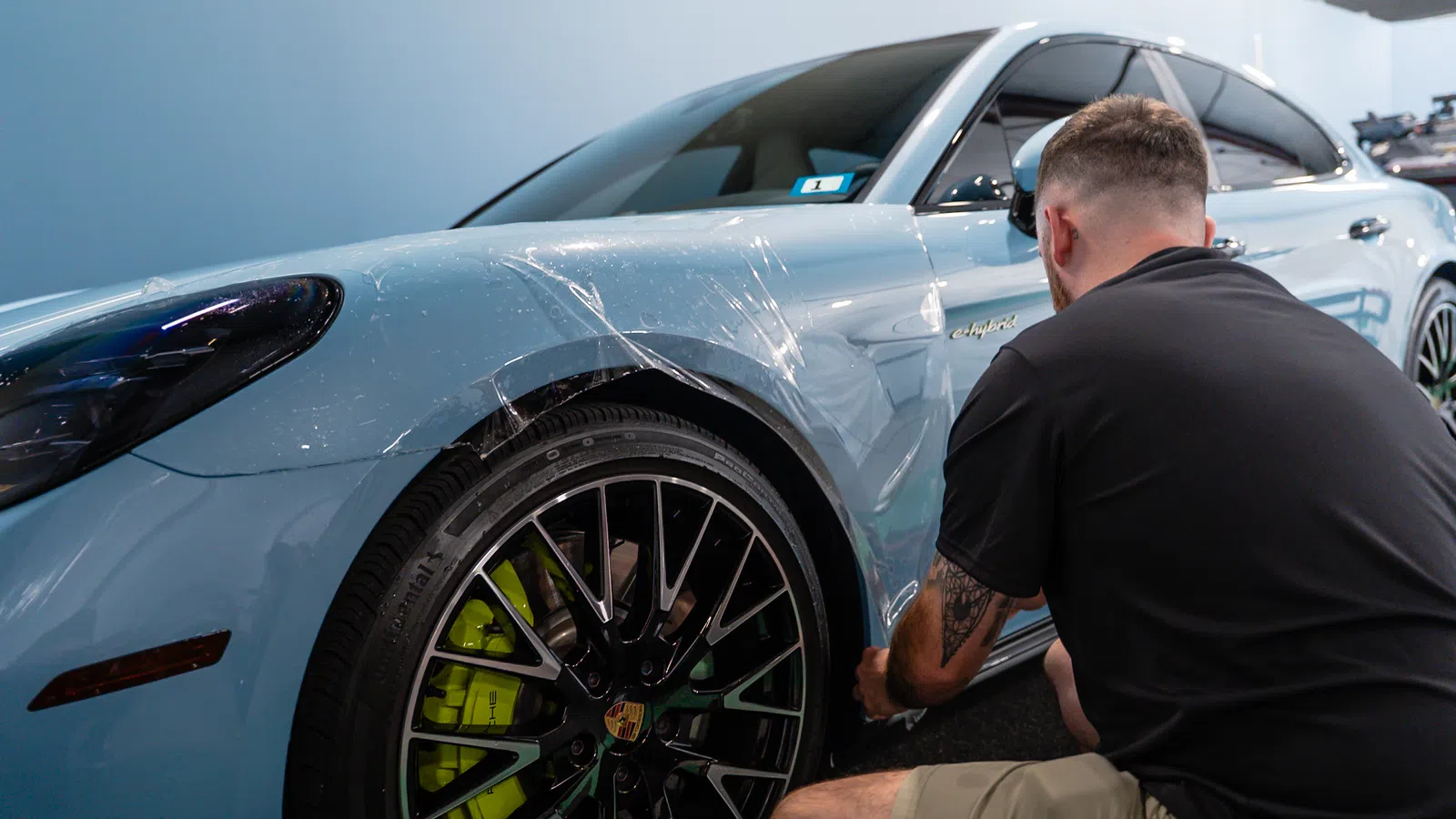 Man applying protective film on light blue Porsche car's front left fender and wheel area indoors.