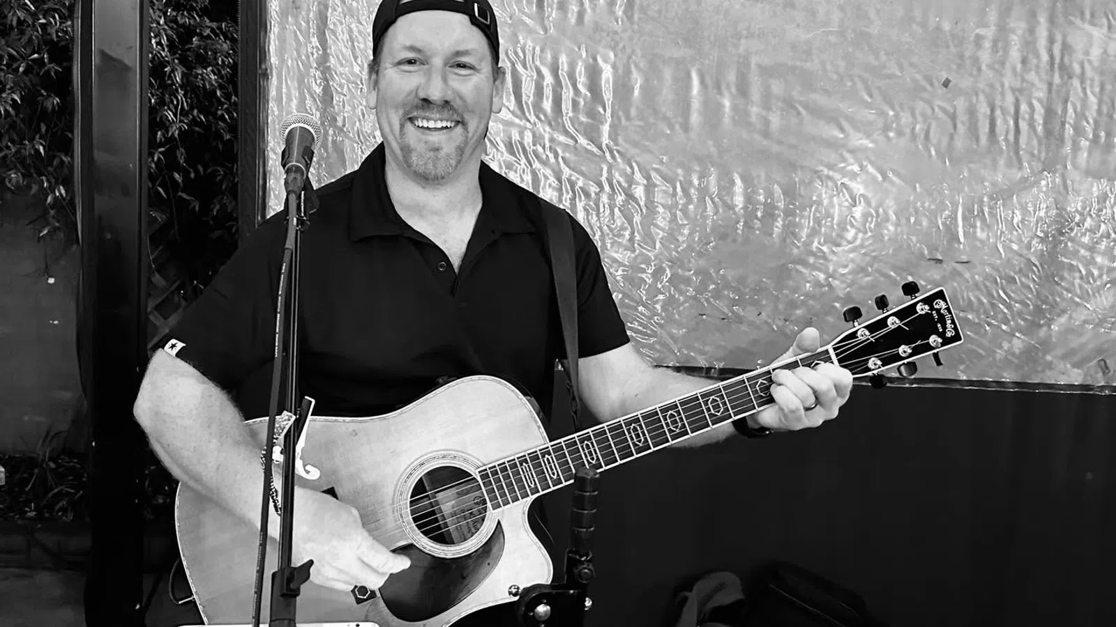 Smiling man wearing a cap plays acoustic guitar and sings into a microphone on stage indoors.