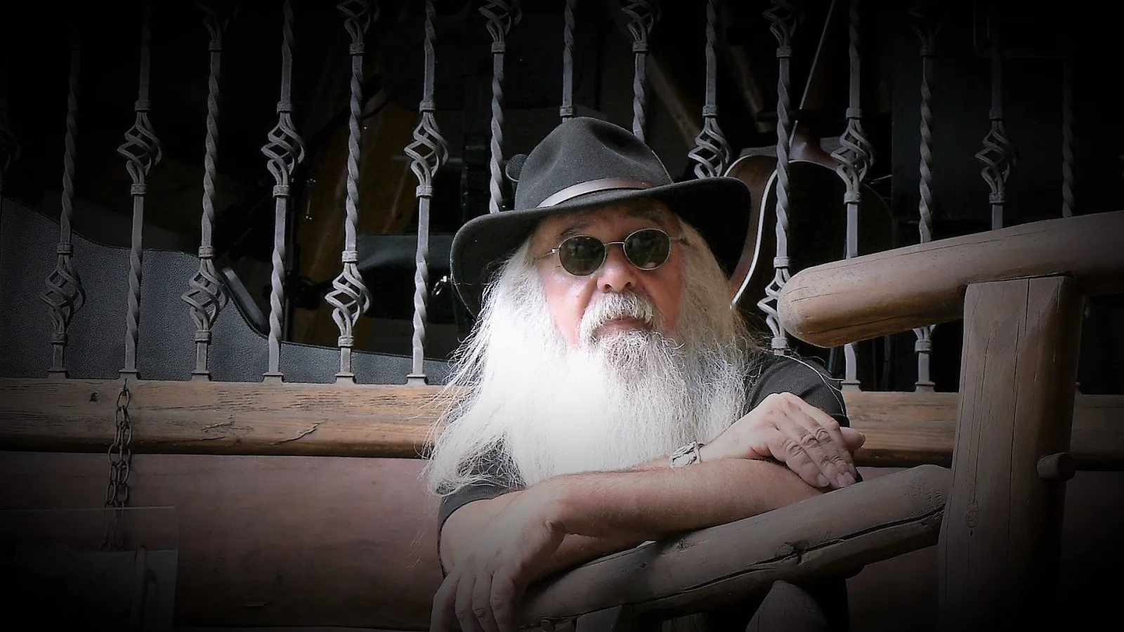 Elderly man with long white beard and sunglasses wearing a black hat leaning on wooden railing indoors.