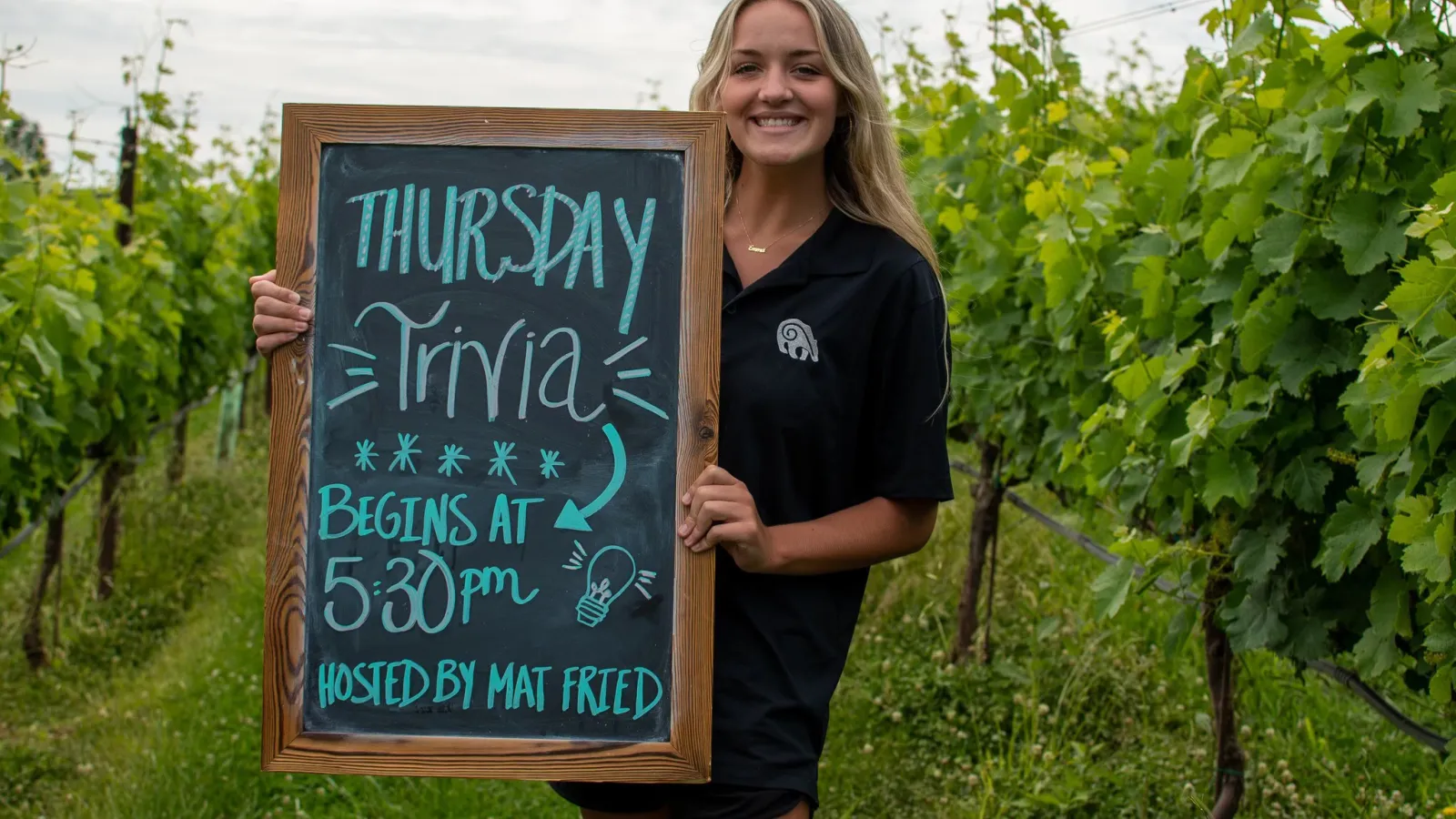 Smiling woman in black shirt holding chalkboard sign for Thursday Trivia at 5:30 pm outdoors in vineyard setting.