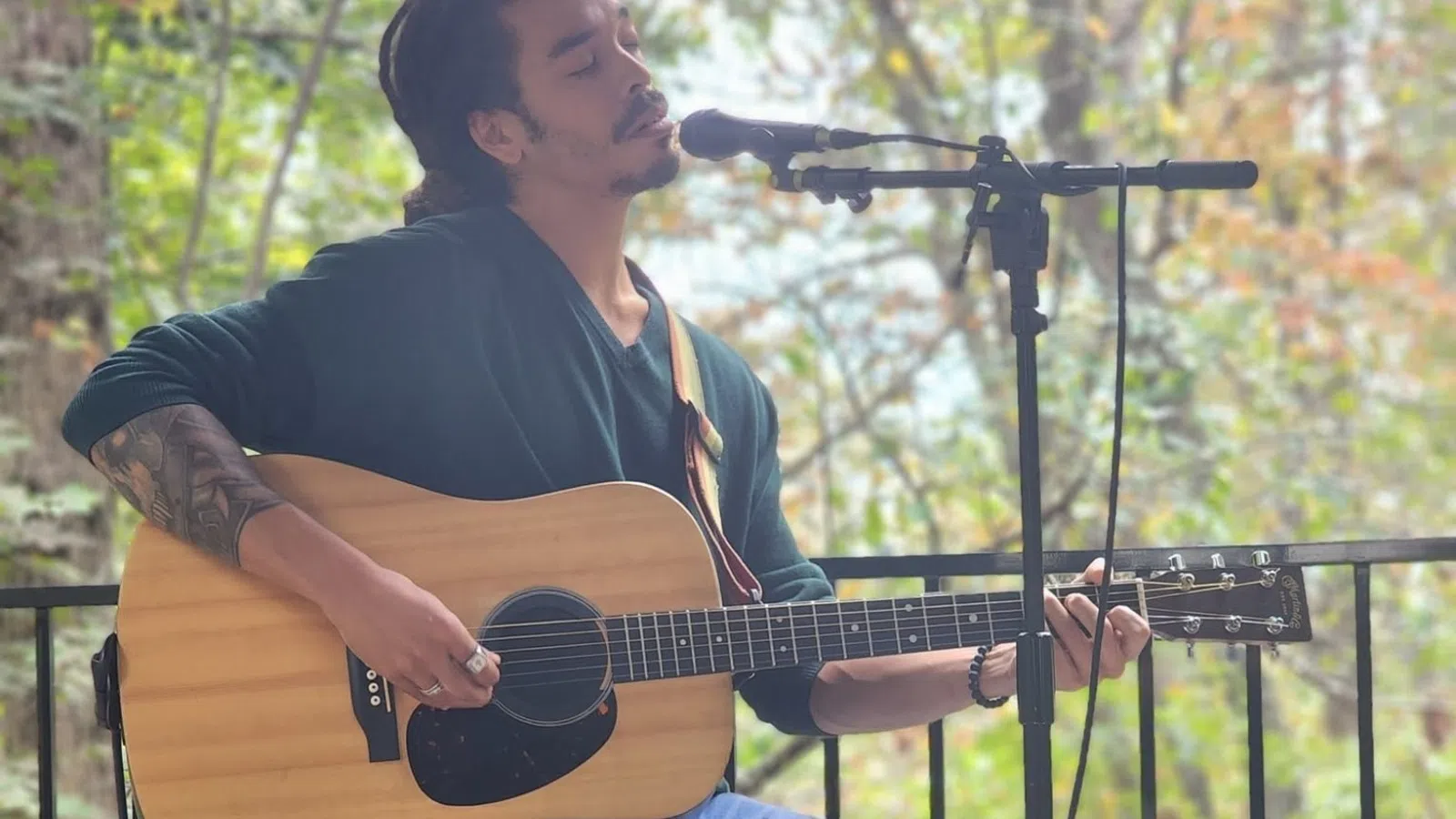 Man playing acoustic guitar and singing into microphone outdoors on wooden deck surrounded by trees.