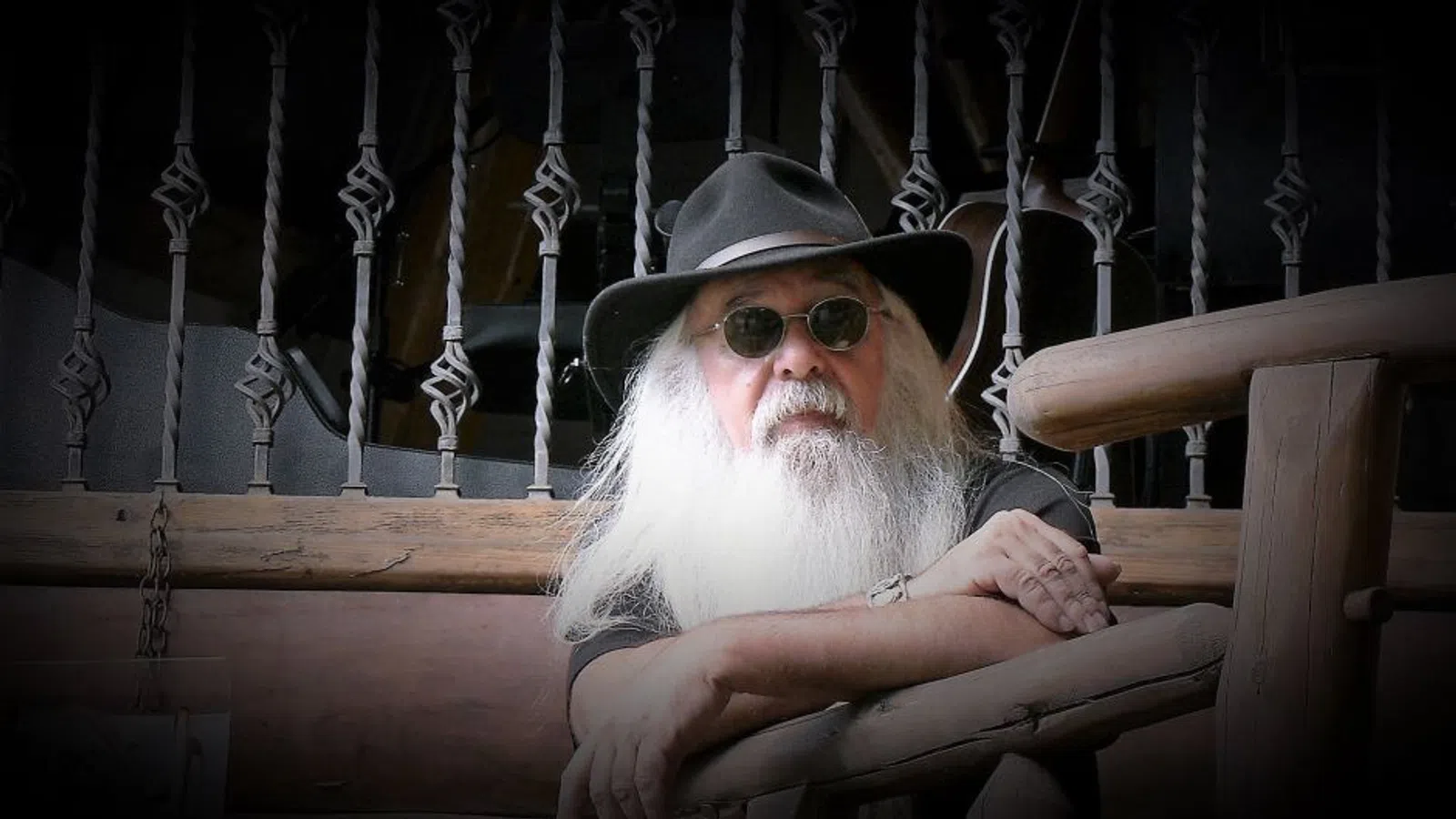 Elderly man with long white beard and sunglasses wearing a black hat, leaning on wooden railing indoors.