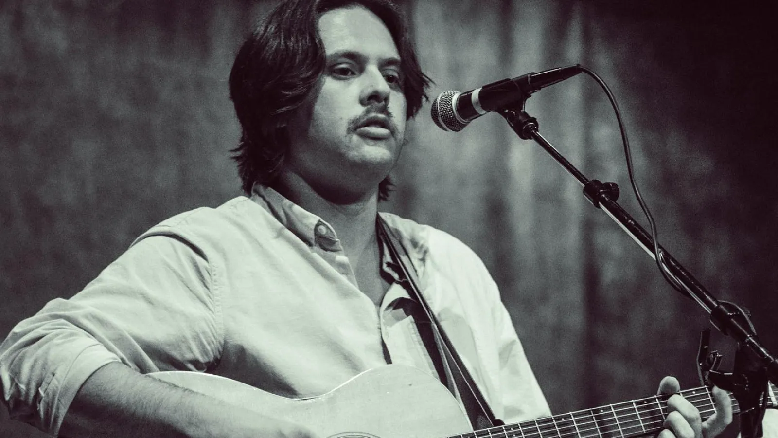 Male musician playing acoustic guitar and singing into microphone on stage in black and white photo
