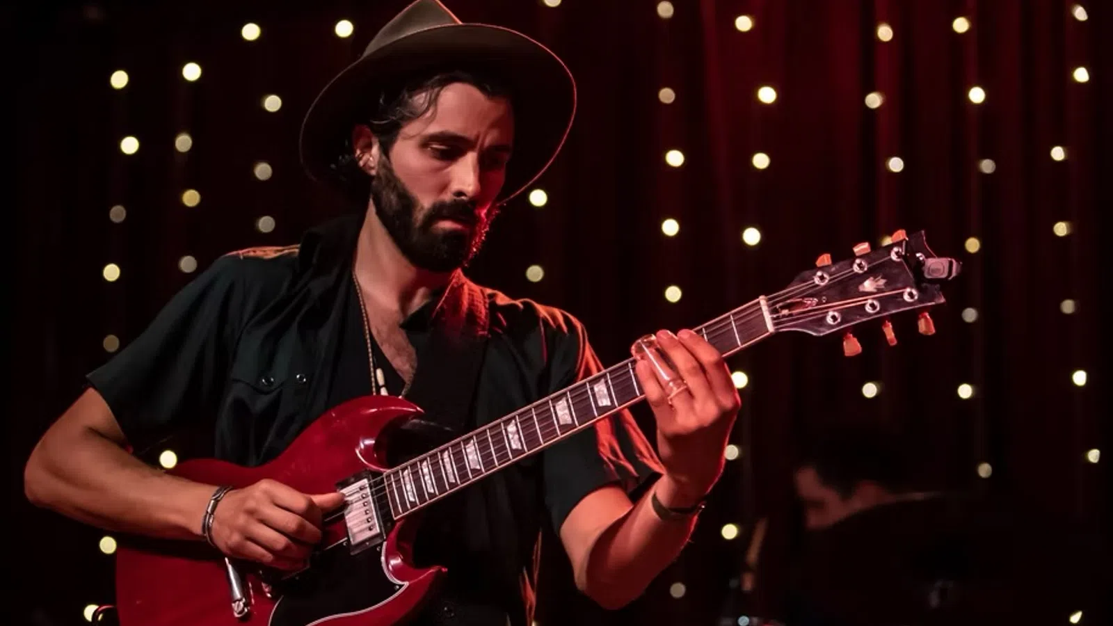 Bearded man in hat playing red electric guitar on stage with string lights background.