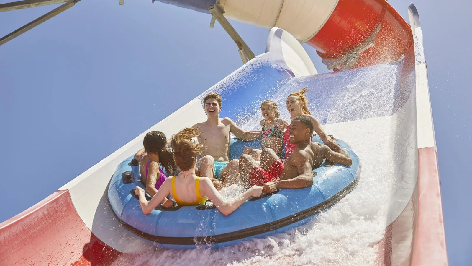 Group of kids and teenagers riding a blue inflatable raft down a colorful water slide on a sunny day.
