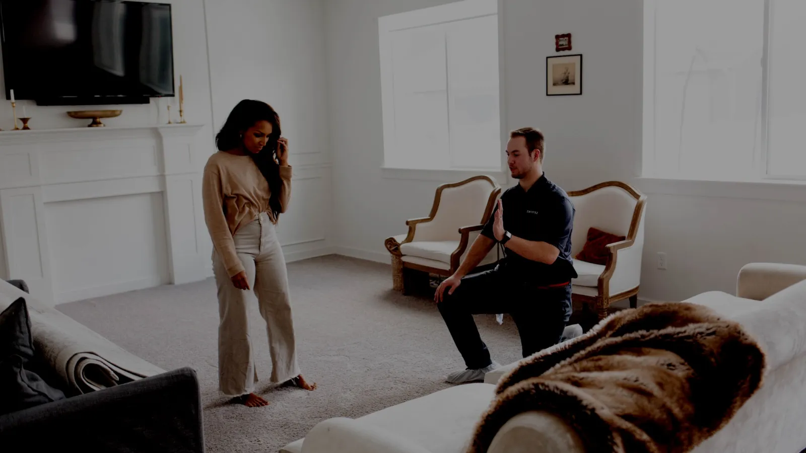 Technician kneeling in living room explaining repair to woman standing barefoot on carpet near couches