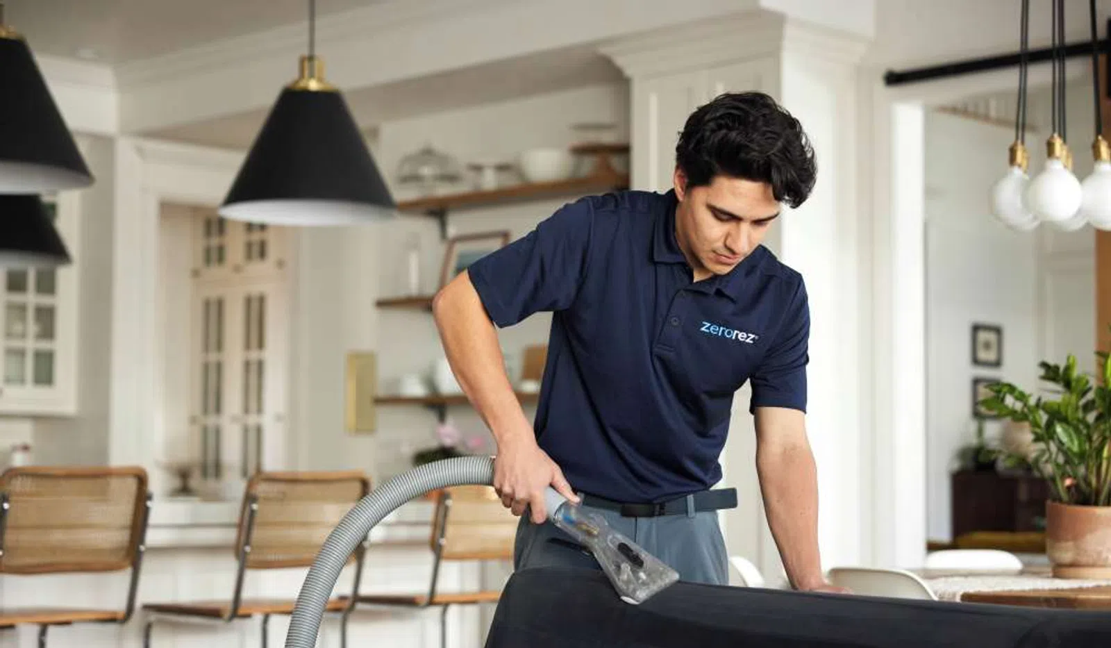 Professional cleaner using a vacuum to clean a black upholstered sofa in a modern living room.