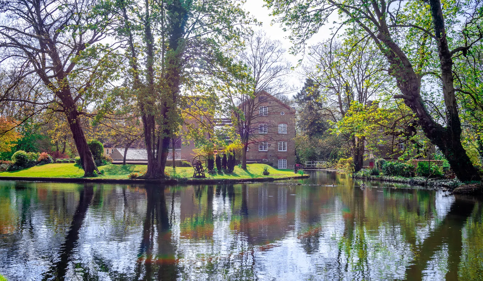 Serene river scene with trees and a brick building reflecting in calm water on a sunny spring day