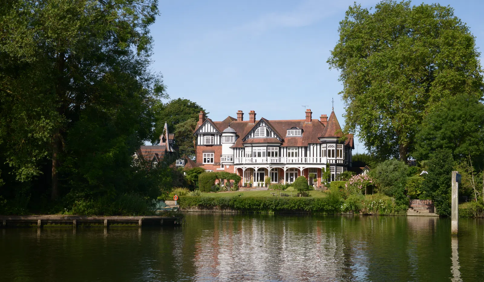 Large Tudor-style house by a river with lush green trees and a clear blue sky reflected in the water.