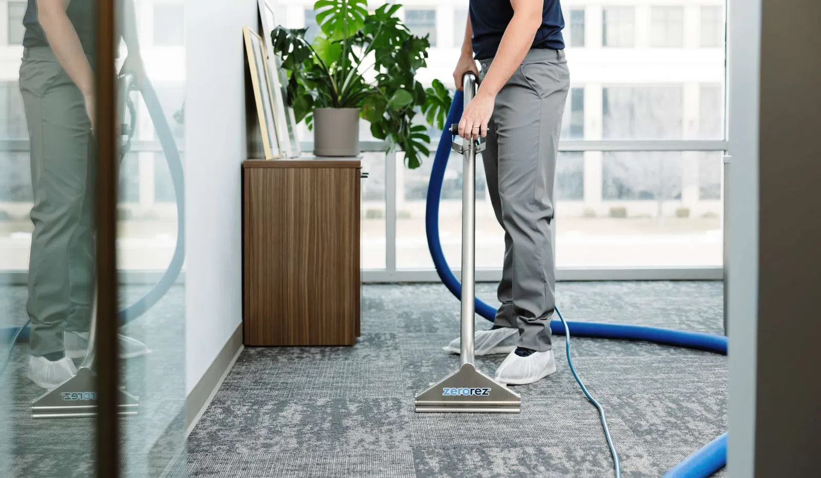 Person cleaning office carpet with Zerorez carpet cleaning machine near plant and wooden cabinet.