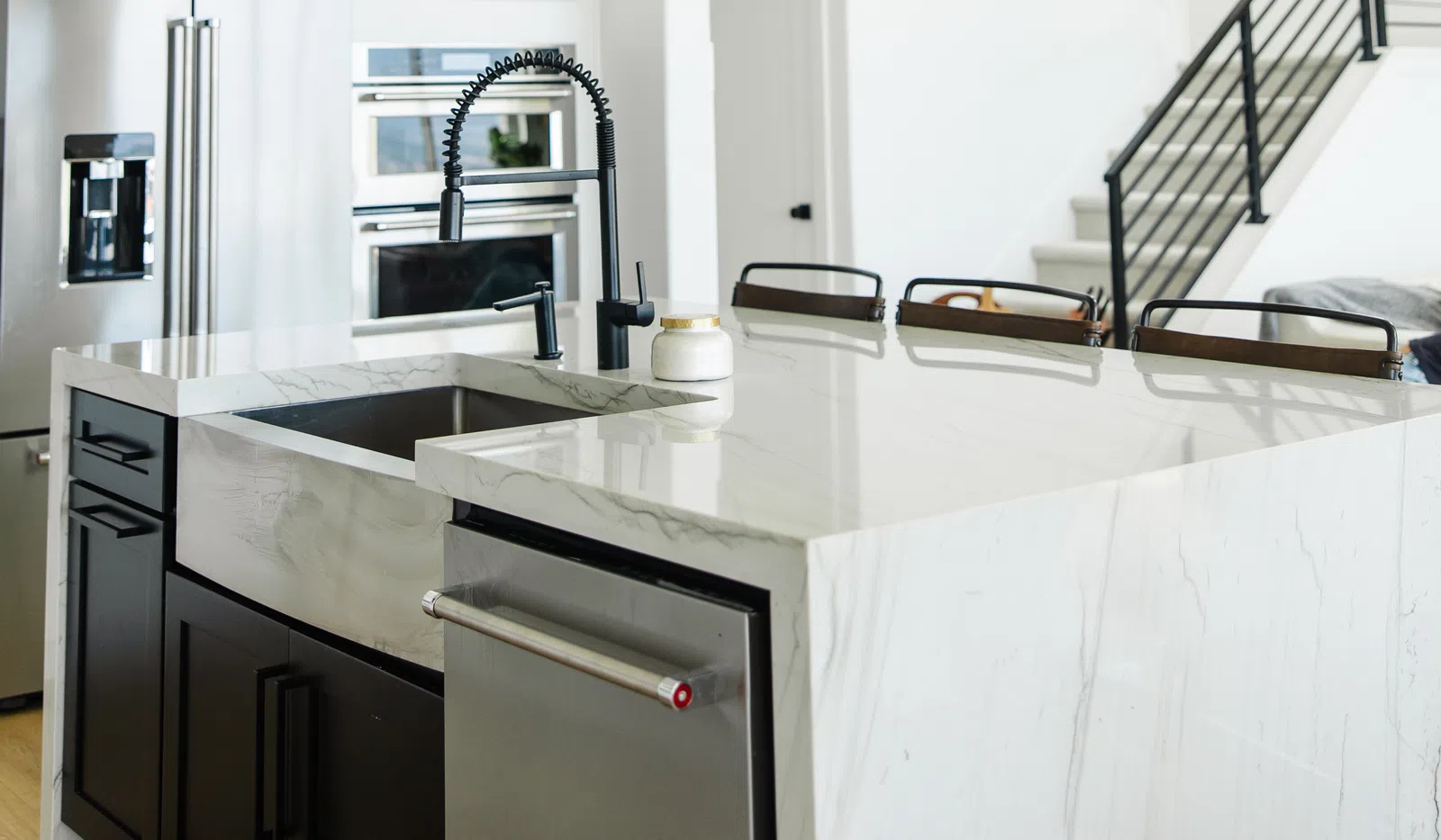 Modern kitchen with marble island, black faucet, pendant lights, stainless steel appliances, and staircase in background.