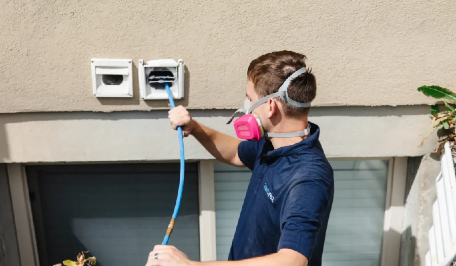 Technician wearing a protective mask cleans an outdoor dryer vent with a blue hose against a beige wall.