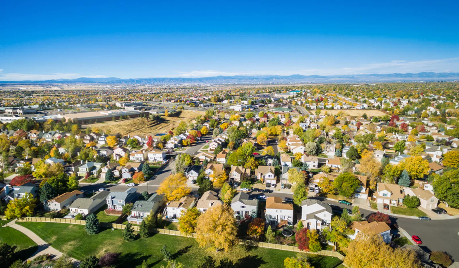 Aerial view of a suburban neighborhood with houses, autumn trees, and distant mountains under a clear blue sky.