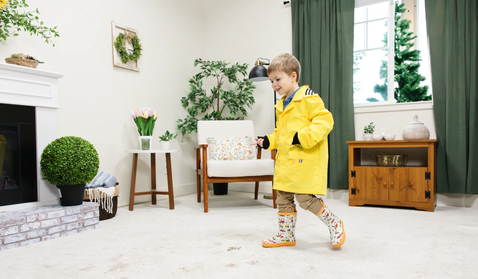Young boy in a yellow raincoat and patterned boots walks on a carpet with muddy footprints in a cozy living room.