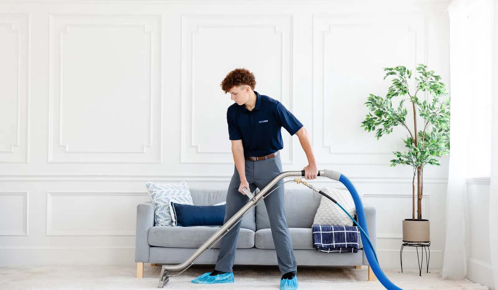 Professional cleaner using carpet cleaning machine in a bright, modern living room with gray sofa and green plant.